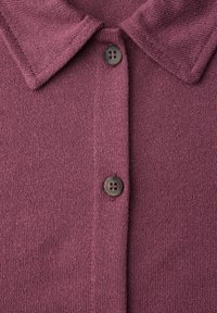 Gros plan sur le col d'une chemise violette douce et tricotée, avec deux boutons foncés attachés sur la patte de boutonnage avant.