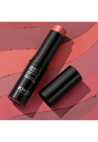 Svart cylindrisk krämig rougepenna med en rosa topp, märkt "Velvet Touch", liggande på en texturerad bakgrund i rosa nyanser.