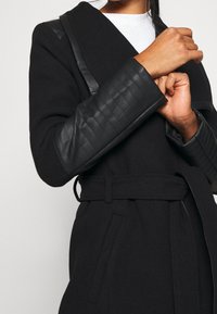 Abrigo negro con diseño envolvente, que cuenta con mangas de cuero negro acolchado y un cinturón a juego. Textura suave con cuello inclinado y bolsillos laterales.