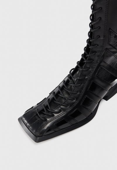 Black leather lace-up boot with a square toe, featuring shiny and matte textures, cut-out rectangular accents along the sides, and a thick sole.