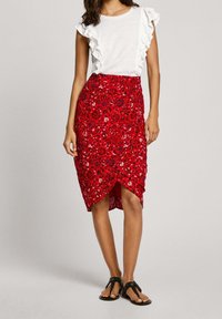 Roter floral gemusterter Wickelrock mit einem hohen Schlitz, kombiniert mit einem weißen rüschengeschmückten Kurzarmoberteil. Das Model trägt außerdem schwarze Sandalen.
