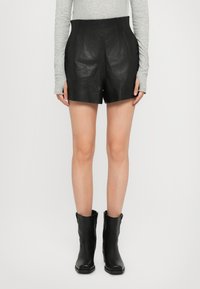 Persona che indossa pantaloni corti in pelle nera a vita alta, maglione grigio a maniche lunghe con buchi per il pollice e stivaletti neri alla caviglia, in piedi su uno sfondo bianco.