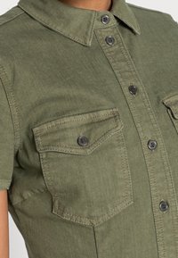 Primo piano di una camicia a maniche corte verde oliva con tasche sul petto e bottoni neri sulla pattina frontale e sui lembi delle tasche.