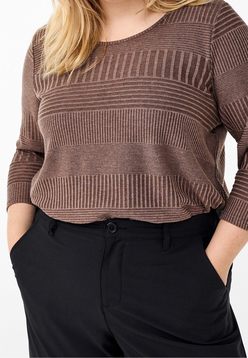 Femme portant une chemise marron rayée à manches longues rentrée dans un pantalon noir taille haute, les mains dans les poches.
