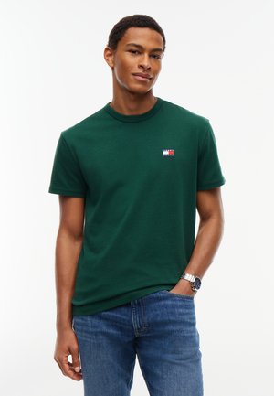 Jeune homme portant un tee-shirt vert à manches courtes avec un petit logo sur la poitrine et un jean bleu, debout avec une main dans la poche.