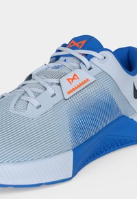 Λευκό και μπλε αθλητικό παπούτσι με υφασμάτινες πλευρές με υφή, λευκά κορδόνια και πορτοκαλί λογότυπο στη γλώσσα και στην πλευρική ετικέτα.