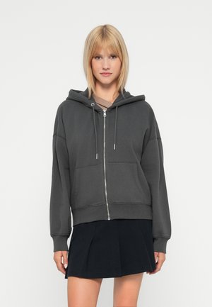 Haut de sweat à capuche zippé gris en tissu doux, avec une grande capuche, des poches avant et une coupe décontractée, porté sur un haut marron et associé à une jupe noire.