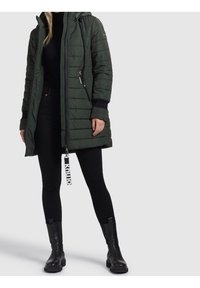 Długi zielony puchowy płaszcz z kapturem, zapinany na zamek, z elastycznymi mankietami i wyraźną metką z logo. Zestawiony z czarnymi legginsami i botkami za kostkę.