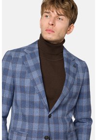 Niebieska marynarka w kratę, wykonana z teksturowanej tkaniny, z brązowym swetrem pod spodem. Posiada klapy o wycięciu i zapięcie na jeden guzik.