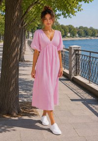 Femme en robe rose et baskets blanches marchant le long d’un chemin bordé d’arbres au bord de l’eau par une journée ensoleillée.