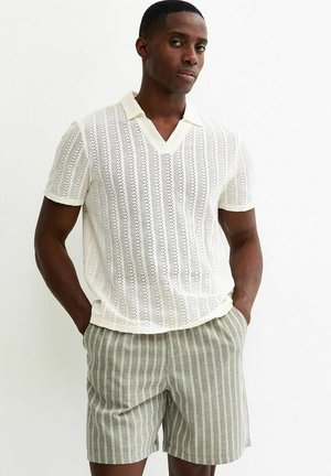 Uomo che indossa una maglietta bianca a maniche corte in maglia con colletto e pantaloncini verde chiaro con strisce verticali bianche, in piedi con le mani nelle tasche.