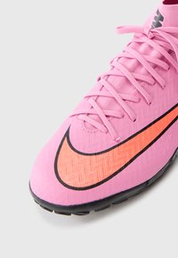 Pink soccer shoe with textured material, black and bright orange Nike swoosh, lace-up front, and a black rubber sole.