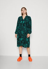 Vihreä-mustakuvioinen mekko, pitkät hihat, V-aukko, rento istuvuus, polvenpituinen, yhdistetty kirkkaan oransseihin kenkiin ja mustiin sukkiin.