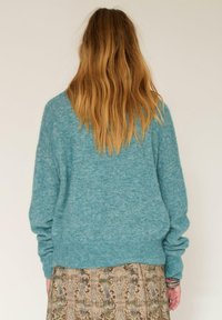 Pull en laine turquoise avec une coupe décontractée, épaules tombantes et ourlet côtelé, associé à une jupe à motifs présentant des tons terre.