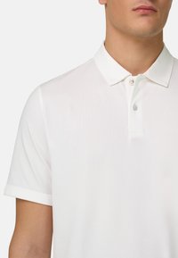 Homme portant un polo blanc à manches courtes avec un col et deux boutons, sur un fond clair uni.