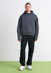Man staat op een groene vloer, draagt een donkergrijze oversized hoodie, zwarte broek en witte sneakers, tegen een effen witte achtergrond.