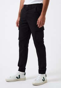 Pantalones cargo negros con bolsillos laterales, corte ajustado y tejido trenzado. Combinados con zapatillas blancas con acentos verdes y puños acanalados.