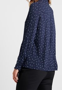 Blusa blu navy con motivo di ancore, tessuto leggero, maniche lunghe, polsino con bottone, dettagli sul retro e vestibilità rilassata.