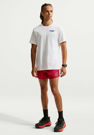 Ung mand med fletninger iført en hvid t-shirt, røde atletikshorts, sorte strømper og sorte løbesko med røde såler, stående mod en hvid baggrund.
