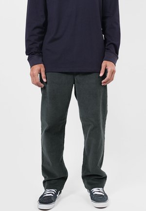 Homme portant un pantalon en velours côtelé vert foncé, une chemise à manches longues bleu marine et des baskets noires et blanches, debout sur un fond blanc.