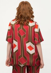 Rotes Kurzarmhemd aus einem glänzenden Stoff mit einem geometrischen Muster aus braunen, orangefarbenen und cremefarbenen Formen, in lässigem, lockeren Schnitt.