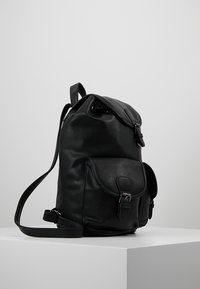 Czarna skórzana plecak z zaokrąglonym szczytem, regulowanym paskiem, przednią kieszenią i detalami w postaci klamry. Gładka tekstura, minimalne szwy i brak widocznych wzorów.