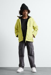 Veste isolante jaune avec des accents noirs, T-shirt noir, pantalon cargo gris et baskets blanches, portée avec un bonnet noir.
