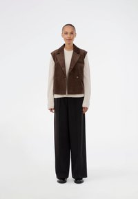 Brązowy sztruksowy bezrękawnik na kremowym swetrze, połączony z szerokimi czarnymi spodniami i czarnymi butami. Gładkie tło.