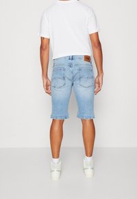 Ljusblå denimshorts med rak skärning, fem fickor och en brun läderlogga vid midjan, kombinerat med vita sneakers.