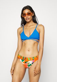 Top de biquíni azul com decote em V e alças cruzadas; parte de baixo laranja com padrão floral e cintura com franzido. Óculos de sol transparentes laranja.