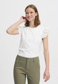 T-shirt en coton blanc avec des manches en dentelle à œillets, associé à un pantalon ajusté vert olive. Encolure ronde simple, texture lisse.