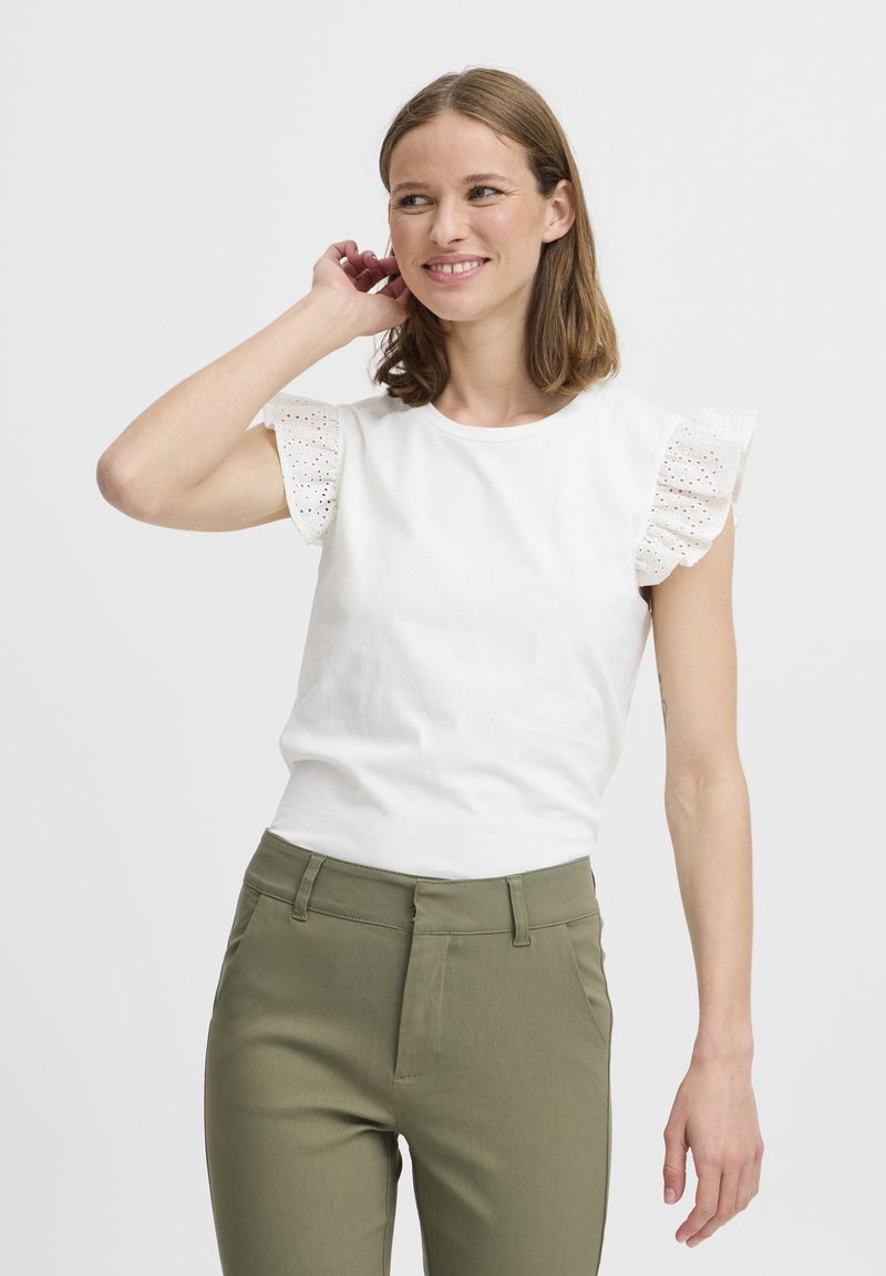 T-shirt en coton blanc avec des manches en dentelle à œillets, associé à un pantalon ajusté vert olive. Encolure ronde simple, texture lisse.