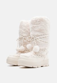 White faux fur boots with a tall shaft, accented by cord laces and pom-poms. Thick rubber soles provide traction and durability.