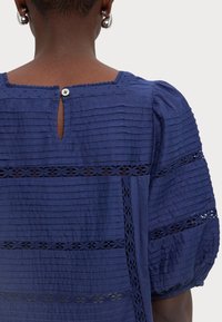 Top blu navy con pieghe orizzontali, dettagli in pizzo, scollo tondo con chiusura a bottone e maniche a sbuffo. Tessuto strutturato.