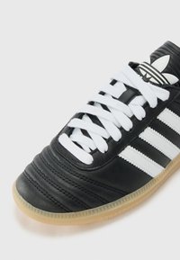 Baskets en cuir noir avec lacets blancs et trois bandes blanches. La semelle en caoutchouc beige présente des détails texturés. Marque argentée sur la languette.