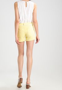 Donna che indossa una blusa senza maniche bianca, pantaloni corti gialli con una cintura bianca e mule con tacco quadro bianchi, in piedi girata di spalle su uno sfondo semplice.