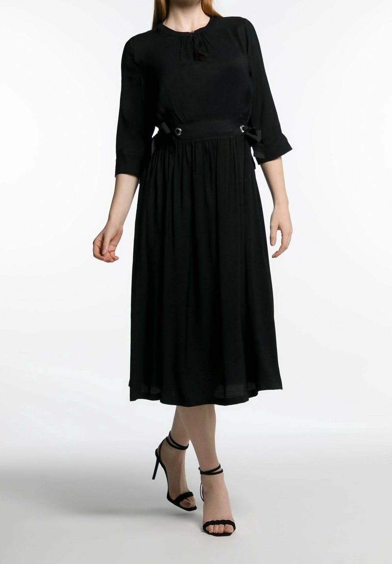 Svart midiklänning med rund halsringning, trekvartsärmar och sidoknytningar med metalliska detaljer, tillverkad av lättviktsmaterial.