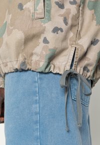 Kamuflage-mønstret letvægtsjakke i beige og grønne nuancer, med elastisk kant og grå snøre; parret med lyseblå denimjeans.