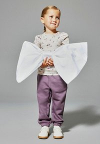 Junges Mädchen mit geflochtenem Haar hält eine übergroße weiße Schleife, trägt ein blumiges Oberteil, lila Hose und weiße Turnschuhe und steht vor einem grauen Hintergrund.