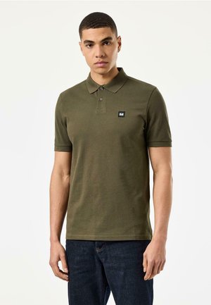 Olivenfarget polo-skjorte i bomullsblanding, med krage og tre knapper, og med en liten firkantet logo på brystet. Båret med mørkeblå jeans.