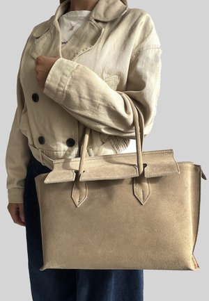Sac à main en suède beige avec une forme structurée, un dessus plat, deux poignées et un rabat avec des accents pointus. Porté par une personne en veste légère.