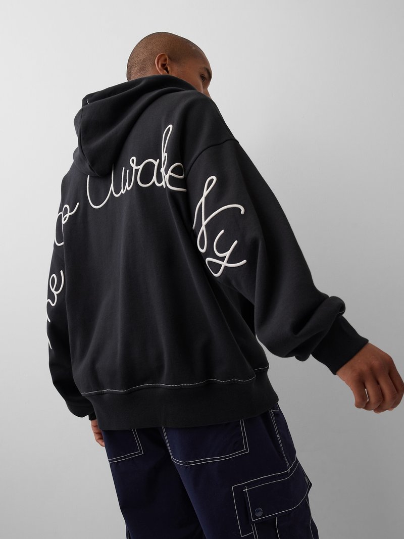 Personne portant un sweat à capuche noir avec un texte en cursive blanc dans le dos et un pantalon cargo bleu marine avec des coutures blanches.