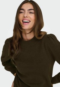 Jeune femme aux longs cheveux bruns portant un pull vert foncé, souriante et regardant légèrement sur le côté, sur un fond uni.