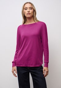 Fuchsia Strickpullover mit Rundhalsausschnitt, langen Ärmeln und gerippten Bündchen. Kombiniert mit dunklen Denim-Hosen. Glatte Textur.