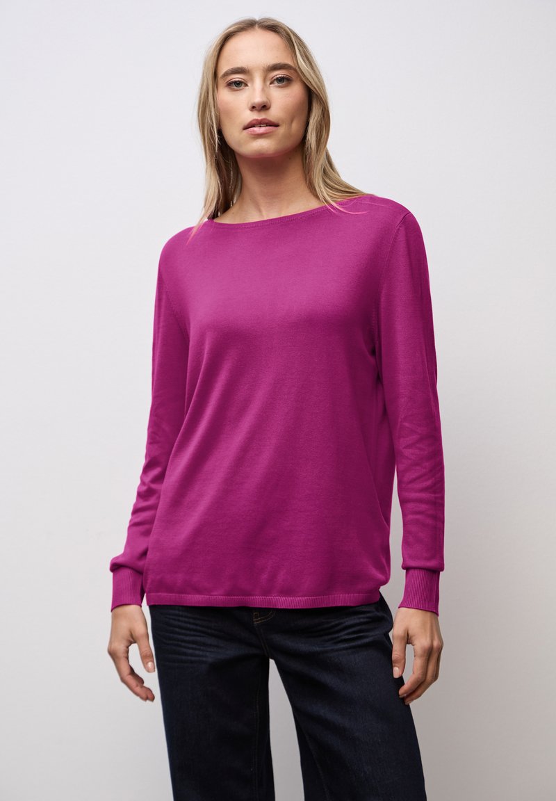 Fuchsia Strickpullover mit Rundhalsausschnitt, langen Ärmeln und gerippten Bündchen. Kombiniert mit dunklen Denim-Hosen. Glatte Textur.