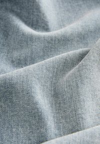 Țesătură din denim albastru deschis, cu o textură netedă, prezentând un model țesut subtil și ușoare unduiri, indicând flexibilitate și moliciune.