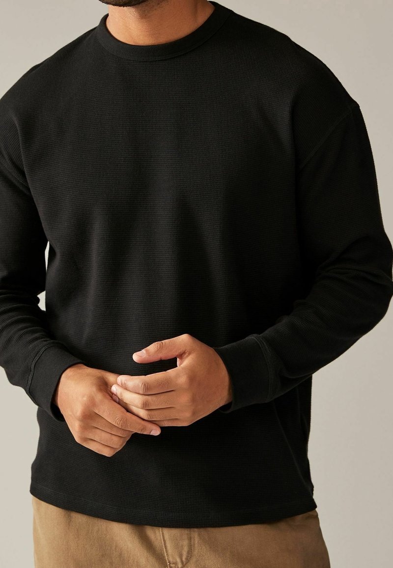 Sweat-shirt noir à manches longues en maille gaufrée, avec col rond et poignets côtes. Texture douce, coupe ajustée, sans logos ni motifs visibles.