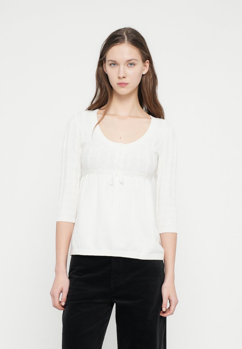 Λευκή πλεκτή μπλούζα με μανίκια τριών τετάρτων, στρογγυλή λαιμόκοψη, λεπτομέρεια με κουμπιά, κορδόνι στη μέση και υφασμάτινες ρίγες κατά μήκος των μανικιών.