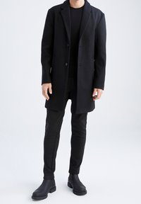 Schwarzer Wollmantel mit Reverskragen und zwei Knöpfen, kombiniert mit enganliegenden schwarzen Hosen und Knöchelstiefeln. Glatte Textur, minimalistisches Design.