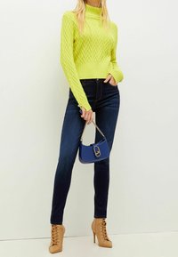 Ljus limegrön stickad tröja med hög hals och zigzag-mönster, i kombination med mörkblå tighta jeans och en liten marinblå handväska.